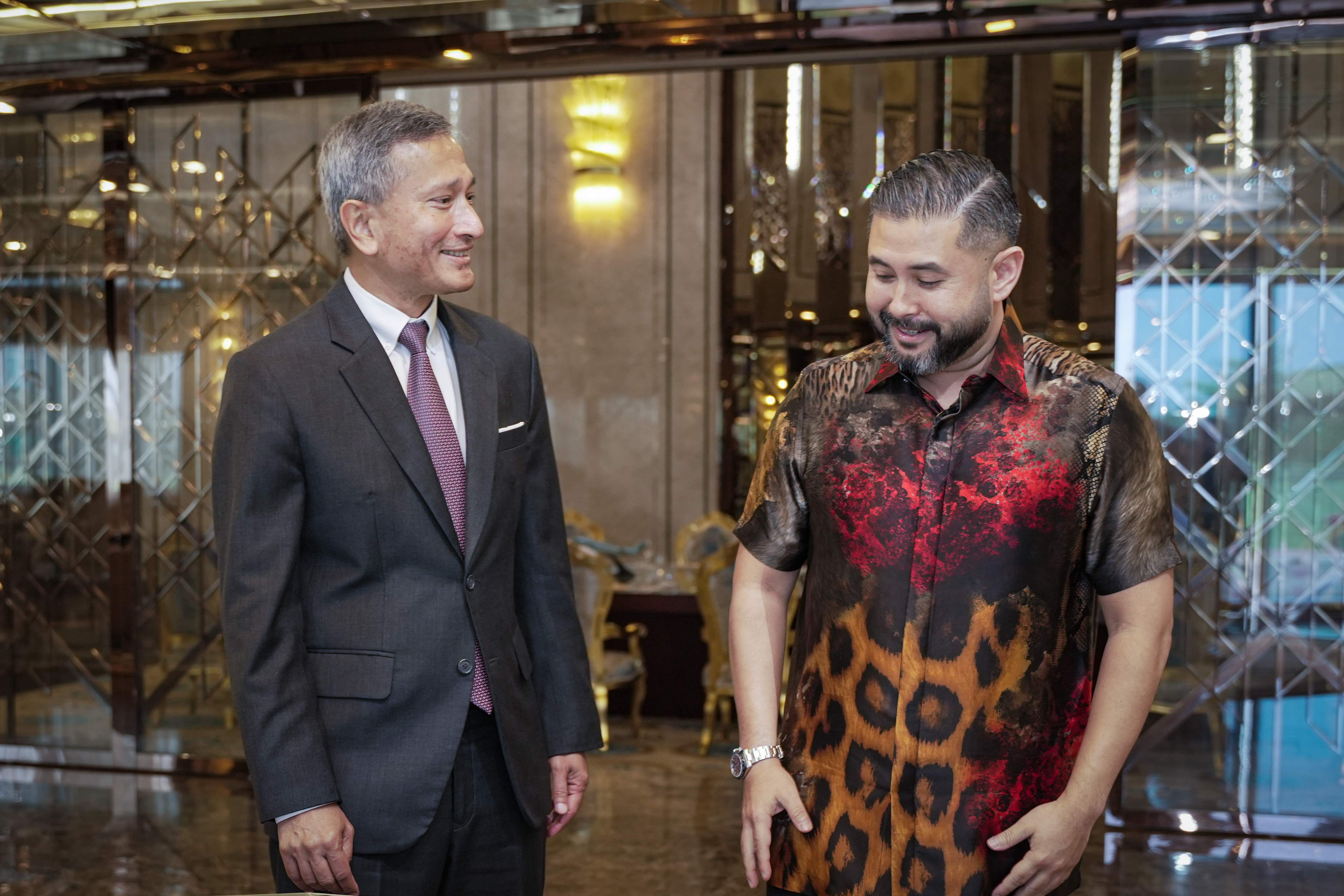Two men stand facing each other. One in a suit and tie, the other in an animal print shirt and wristwatch.